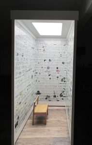 darkness surrounding a light box immersive sculpture of Under the Microscope. It shows the phone booth sized confessional with a small gold chair inside of it inviting a viewer to take a seat and read the walls of text about a mother and child's battle with mental health and substance use and incarceration in Philadelphia and Montgomery County PA Under the Microscope by Maria Maneos